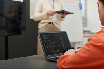Young adult Caucasian woman sitting at desk using laptop with programming code on screen, while middle aged woman standing in background holding tablet during prison education class