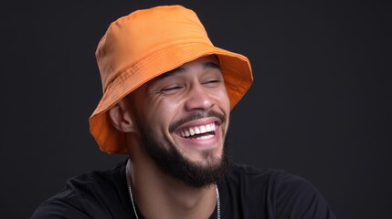 A man sporting an orange bucket hat grinning widely in a brightly lit studio with a black background. 