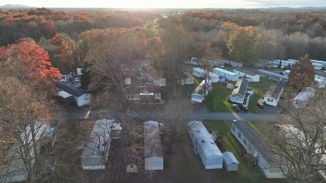Mobile home community in United States during late autumn. Rows of trailer homes surrounded by leafless trees and colorful fall forest, with soft daylight illuminating a quiet residential area. Aerial
