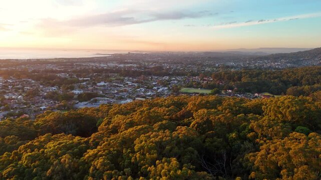 Aerial establishing over forested hills to reveal Illawarra suburbs framed by distant coastline at holden hour