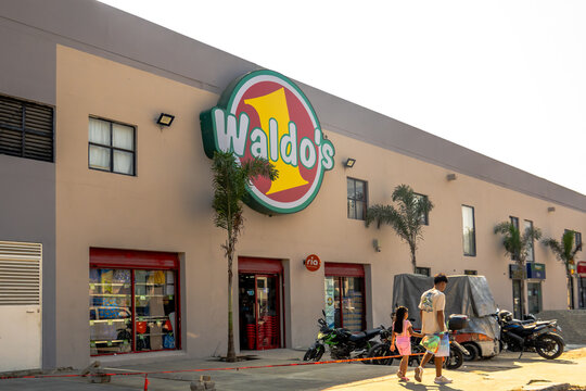Huatulco, Oaxaca, Mexico. Dec 17, 2025. A wide daytime shot of a Waldo's retail building featuring its colorful logo, palm trees, and people walking past parked motorbikes.