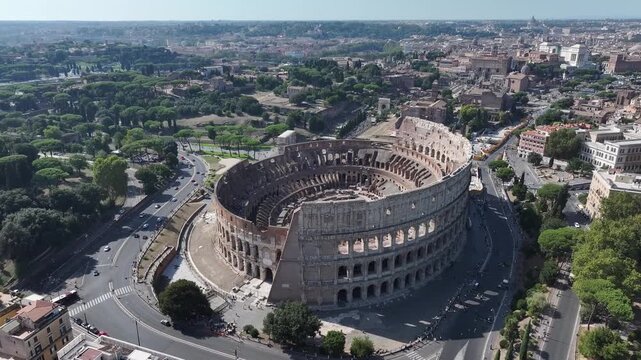 High-angle drone shot over the ancient Colosseum in Rome, Italy, showing the surrounding historical city and bustling road traffic on a clear, sunny day.