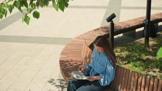 overhead caucasian woman reading on curved brick bench, aerial park composition with leafy frame and geometric pavement, quiet solitary reading moment, denim attire and focused posture, urban design