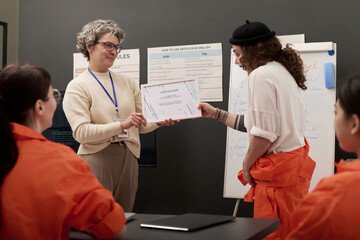 Middle aged Caucasian woman handing certificate to young adult Caucasian woman wearing prison uniform, while two young adult women in orange uniforms sitting at table during prison education class