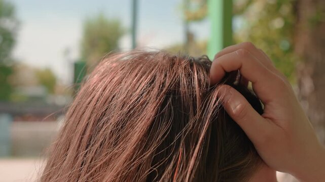 close-up of hand fixing hair at bus stop, sunlit brown strands ruffled by breeze, gentle scalp touch and styling gesture, candid commuter moment with suburban background and relaxed midday light
