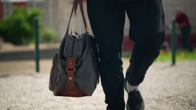 man strolling outdoors, individual with bag walking outdoors, man carrying leather bag in tranquil park setting, casual male figure strolling along park gravel path at dawn with bag swinging