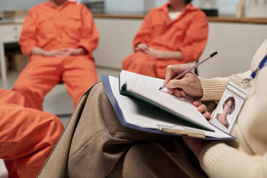 Middle aged Caucasian woman holding clipboard conducting therapy session with group of young adult women in orange prison uniforms sitting in classroom setting
