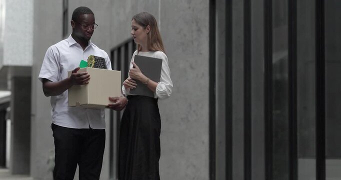 African male employee carries box of office belongings while female coworker encourages him outdoors. Reflecting job loss during an economic downturn and power of workplace support and resilience
