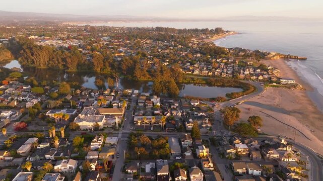 A panning aerial drone shot showing the coastal California towns of Santa Cruz, Aptos, Pleasure Point, and Capitola at sunset with waves gently crashing next to lagoons and beachgoers