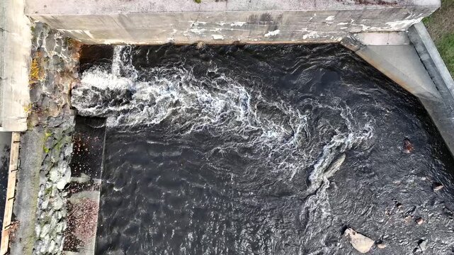 Drone medium top down as water gushes out of culvert pipe from dam into splash pool, static