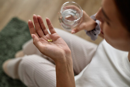 Young adult Caucasian woman sitting and holding two pills in one hand while holding glass of water in other hand, preparing to take medication, partial face visible