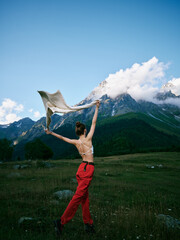 Fototapeta premium Dancer leaping in red pants with scarf in an open meadow beneath towering mountains, wind catching fabric, showcasing movement, freedom, and outdoor adventure