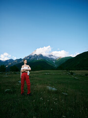 Naklejka premium Woman stands in a mountain meadow with bold redpants, under a clear blue sky, capturing outdoor adventure and serene nature amid alpine scenery