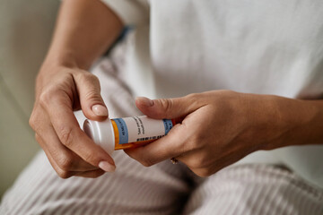 Woman holding prescription medication bottle with both hands, examining label closely, sitting indoors, focus on hands and container