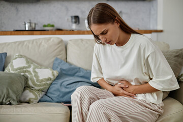 Caucasian young adult woman sitting on sofa holding stomach with both hands appearing to experience...