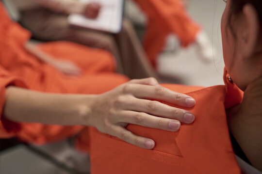 Young adult woman placing hand on shoulder of another young adult woman in orange uniform during prison therapy session, blurred figure with clipboard visible in background