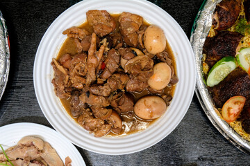 chinese style, Braised pork leg chunks, trotters, and whole boiled eggs in a dark soy-based sauce served on a white ceramic plate. The image is a top-down view on a dark wooden table