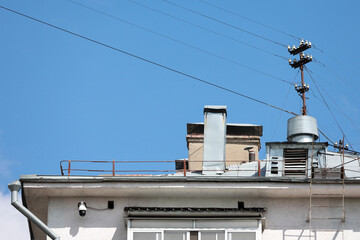 Close Up Of A Building Roof Edge Featuring An Old Metal Antenna, Wires, And A Surveillance Camera Against A Blue Sky.