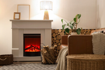 Fototapeta premium Interior of living room with sofa, glowing lamp, wicker table and firewood near fireplace in evening