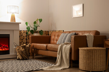 Fototapeta premium Interior of living room with sofa, glowing lamp, wicker table and firewood near fireplace in evening
