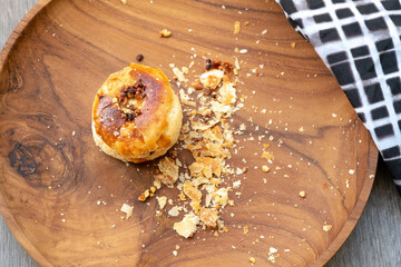 Traditional Indonesian bakpia pastry on a round wooden plate with scattered crumbs. High-angle food photography of a single flaky, golden treat with a checkered cloth detail.
