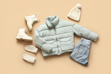 Stylish short puffer jacket with boots, jeans, winter hat and gloves on beige background