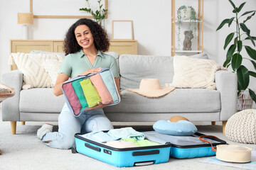 Young African-American woman with organizer packing clothes into suitcase for trip at home