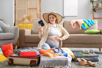 Obraz premium Young pregnant woman with passport, clothes, travelling accessories and full suitcase at home