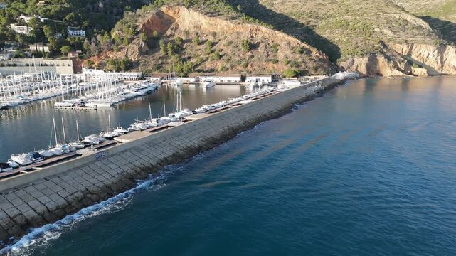 Javea Spain. Aerial drone video of the harbor breakwater at sunset with docked boats, a fishing vessel arriving, and the coastal town backdrop.