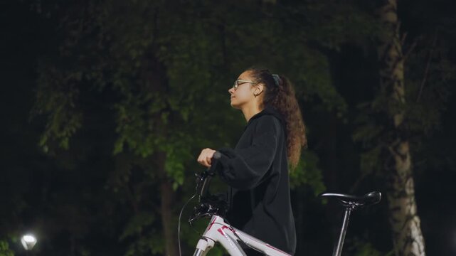 Young Woman Standing Beside Bicycle Under Park Lights At Night, Patient Waiting Pose With Hands On Handlebars, Distant Trees And Soft Glow Create Cinematic Mood, Casual Hoodie Suggesting Commuter