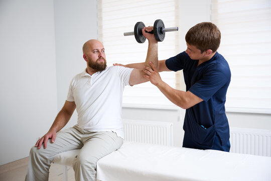 Physiotherapist assisting male patient with shoulder rehabilitation exercise using dumbbell. Man sitting on medical couch while performing physiotherapy workout under supervision of doctor.