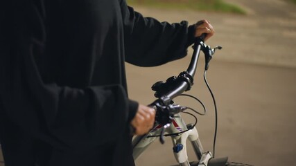 Pavement CloseUp Of Cyclist Holding Handlebars At Night, Person In Loose Black Hoodie Stands Near Curb With Hands On Grips, Ambient Lamplight And Soft Background, Stillness And Reflective Mood