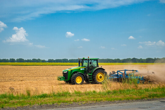 Ukraine, Cherkasy - July 19, 2021:A powerful green tractor with a plow cultivates a vast golden field under a bright blue sky, raising dust. This scene depicts modern agriculture, hard work
