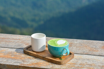 Hot matcha green tea in a blue cup, served with hot water, on a wooden table and blurry nature background.