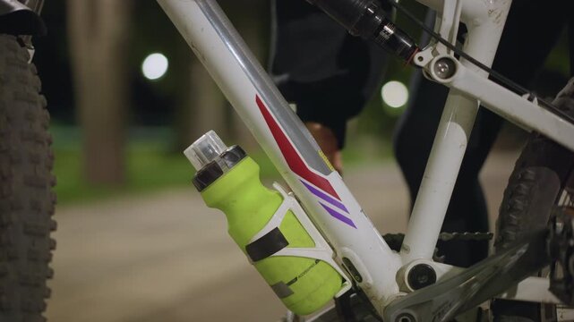 Nighttime Cyclist Pauses For Hydration And Energy, Bicycle Rider Smoothly Grabs Green Bottle During Ride, Cycling Enthusiast Extends Hand For Quick Refill At Nighttime Park Stop Floor