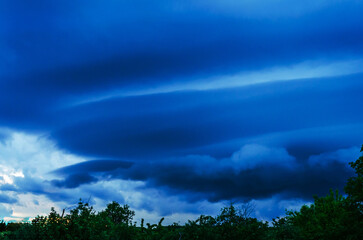 Obraz premium Dramatic dark blue storm clouds gathering ominously over a silhouetted treeline at dusk
