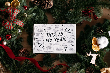 notebook displaying THIS IS MY YEAR resolution list rests on wooden table among festive evergreen...