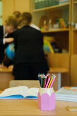 Colorful pencils in a pink holder on a wooden desk, with blurred students collaborating in the background, showcasing the vibrant atmosphere of school education and learning