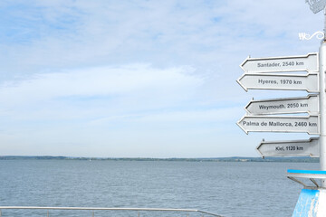 Directional signpost with multiple distance markers pointing towards various destinations over a calm body of water under a clear blue sky, showcasing travel options and exploration