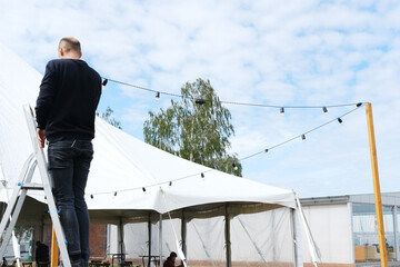 Worker on ladder prepares a large white tent for celebration, hanging decorative lanterns and string lights, creating a festive atmosphere for outdoor event © ribalka yuli