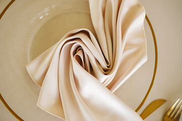 Folded beige napkin on clear glass plate with golden rim and gold fork on beige table surface, arranged elegantly for dining setting with copy space