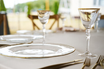 Elegant dining table setting with clear glass plates, crystal wine glasses, and gold-accented cutlery on beige tablecloth with blurred outdoor background and wooden chairs