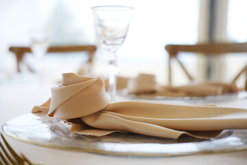 Table setting with beige napkin elegantly arranged on silver plate beside glassware and wooden chairs in bright indoor dining area with soft natural light
