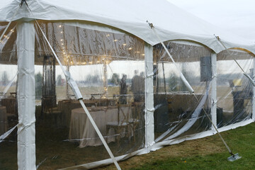 Clear tent structure with white fabric and transparent sides, tables set with white tablecloths inside, outdoor setting with grass and cloudy sky above