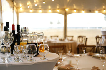 Table setting with wine bottles, glasses, and a pitcher on white tablecloth in a tent with string lights and wooden chairs during an outdoor event