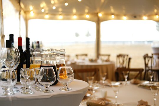 Table setting with wine bottles, glasses, and a pitcher on white tablecloth in a tent with string lights and wooden chairs during an outdoor event