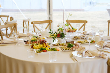 Table set with white tablecloth, plates of food, glasses, cutlery, floral centerpiece, and various dishes arranged on a round table in a tent during an event