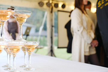 Champagne glasses stacked on white table with sparkling drink inside, blurred people in background, event setting with lighting and equipment visible in tent during daytime
