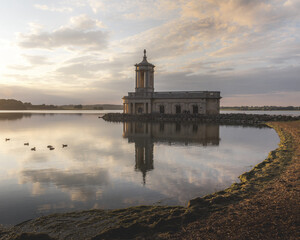 View of Normanton Church's stone facade rises majestically from the tranquil waters, mirroring the soft, pastel sunset, a serene scene, Oakham, England, United Kingdom.