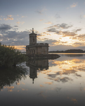 View of Normanton Church stands gracefully on the water's edge, its reflection mirrored in the serene lake under a sky painted with hues of sunset, Oakham, England, United Kingdom.